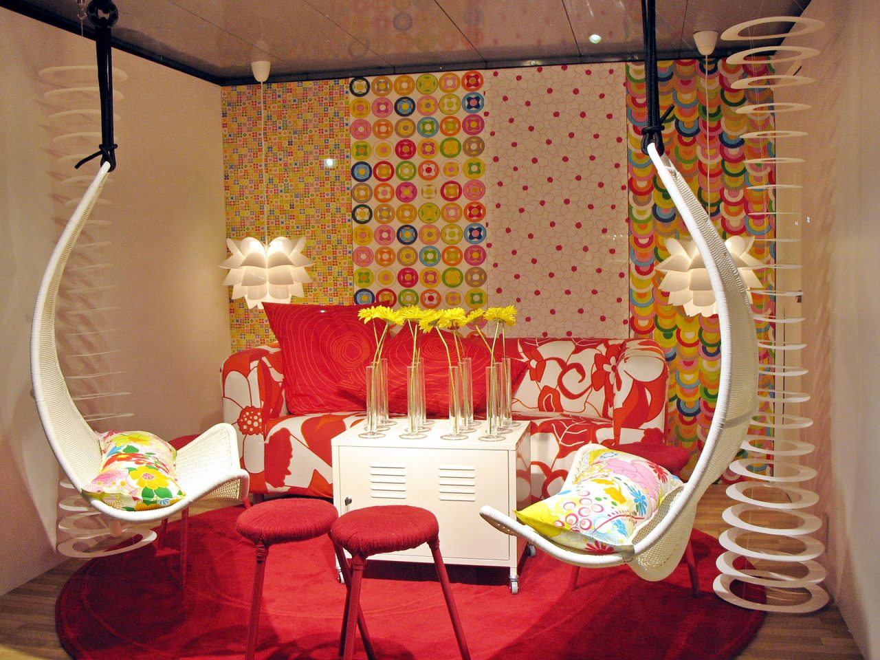 Uppbyggd rumsinteriör i glada färger på liten yta, en liten röd soffa och två vita hängstolar.