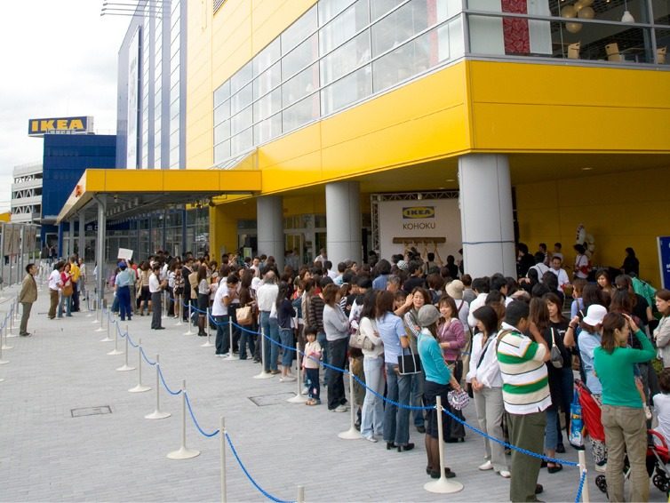 A long queue of dark-haired people winding around a large blue and yellow IKEA store.