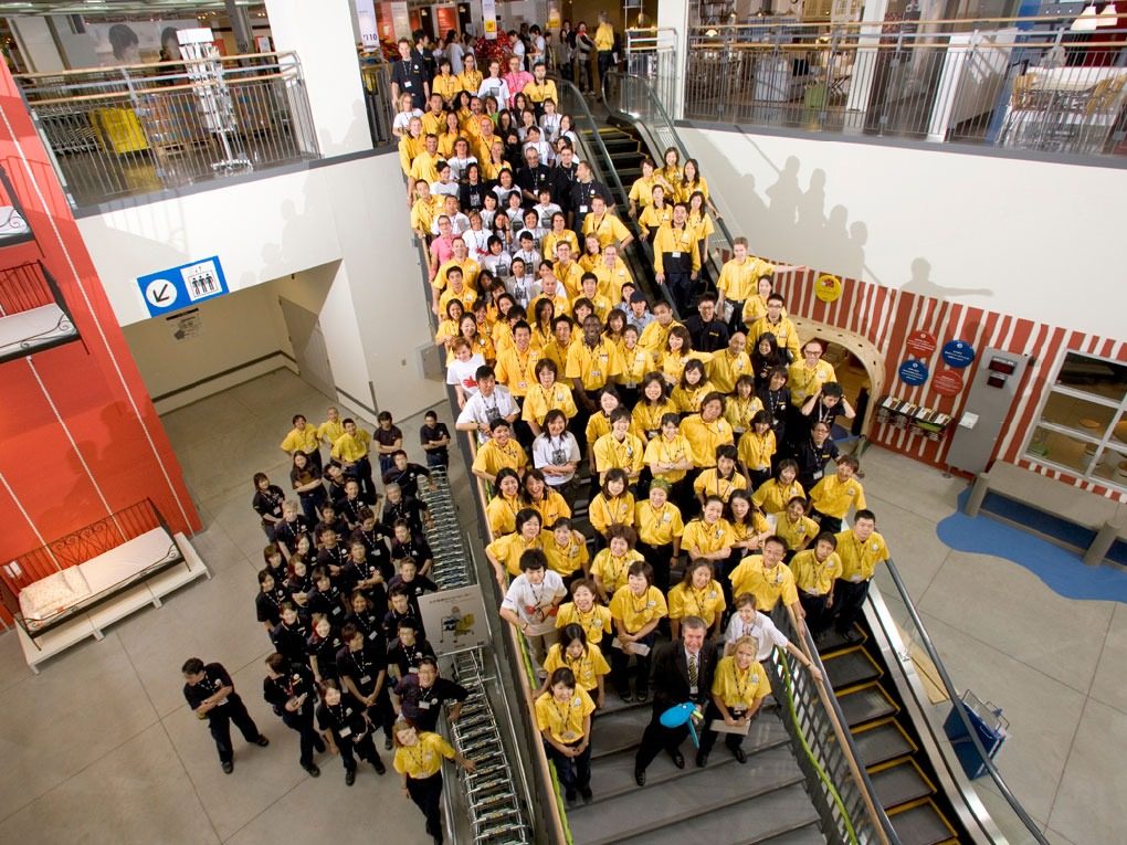 Large group of people in IKEA staff uniforms standing in long staircase and escalator, and on store floor below.