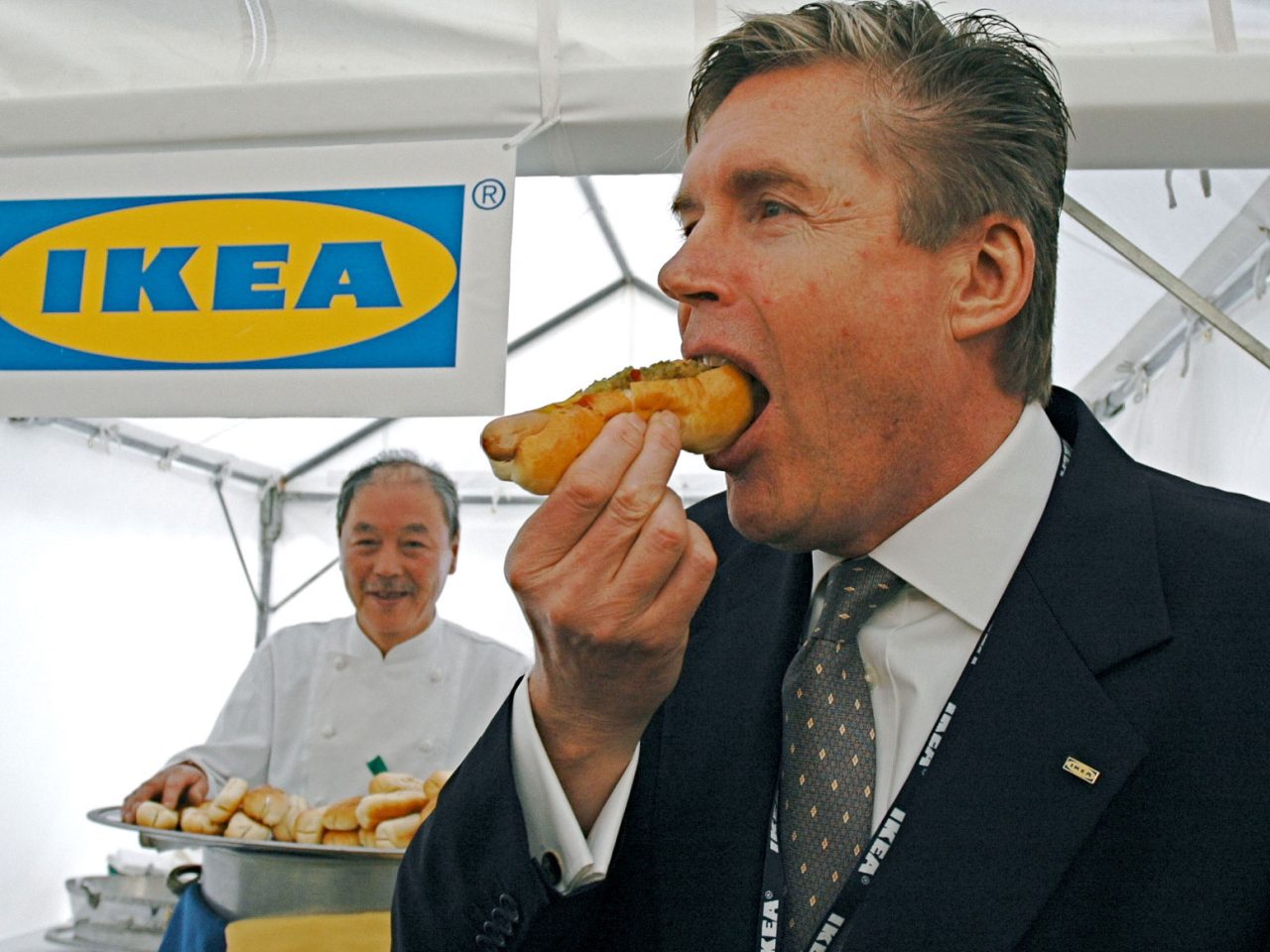 Man in dark suit and tie takes a big bite of a hot dog, in the background a smiling Japanese chef and an IKEA sign.