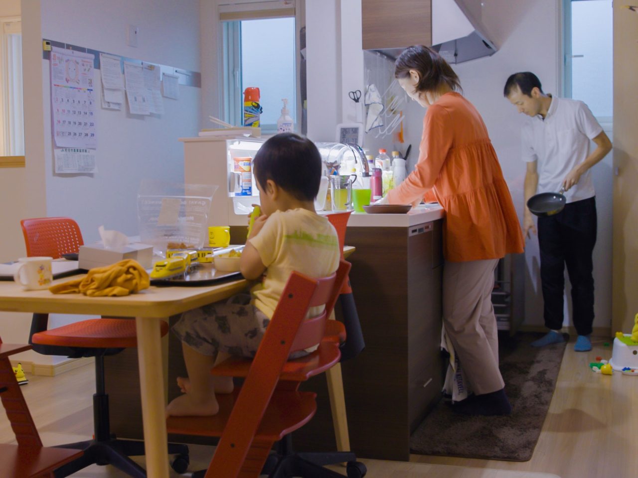 Small child sitting at a dining table in a kitchen, a woman and a man washing and clearing away behind him.