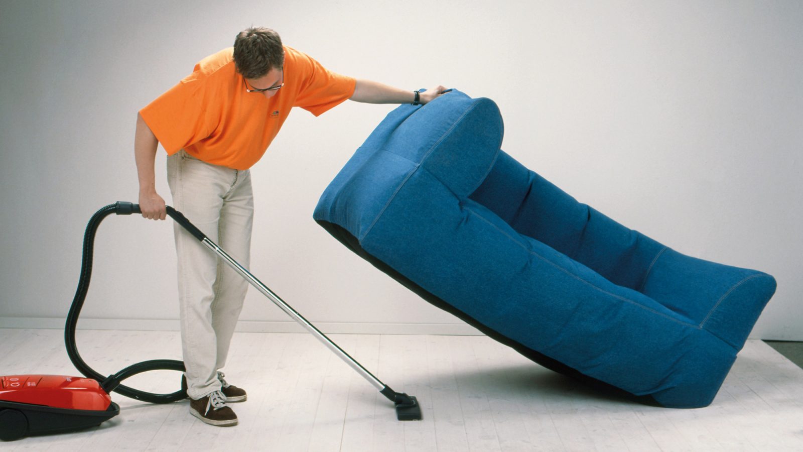 Man in orange T-shirt lifts blue sofa with one hand to vacuum underneath it.