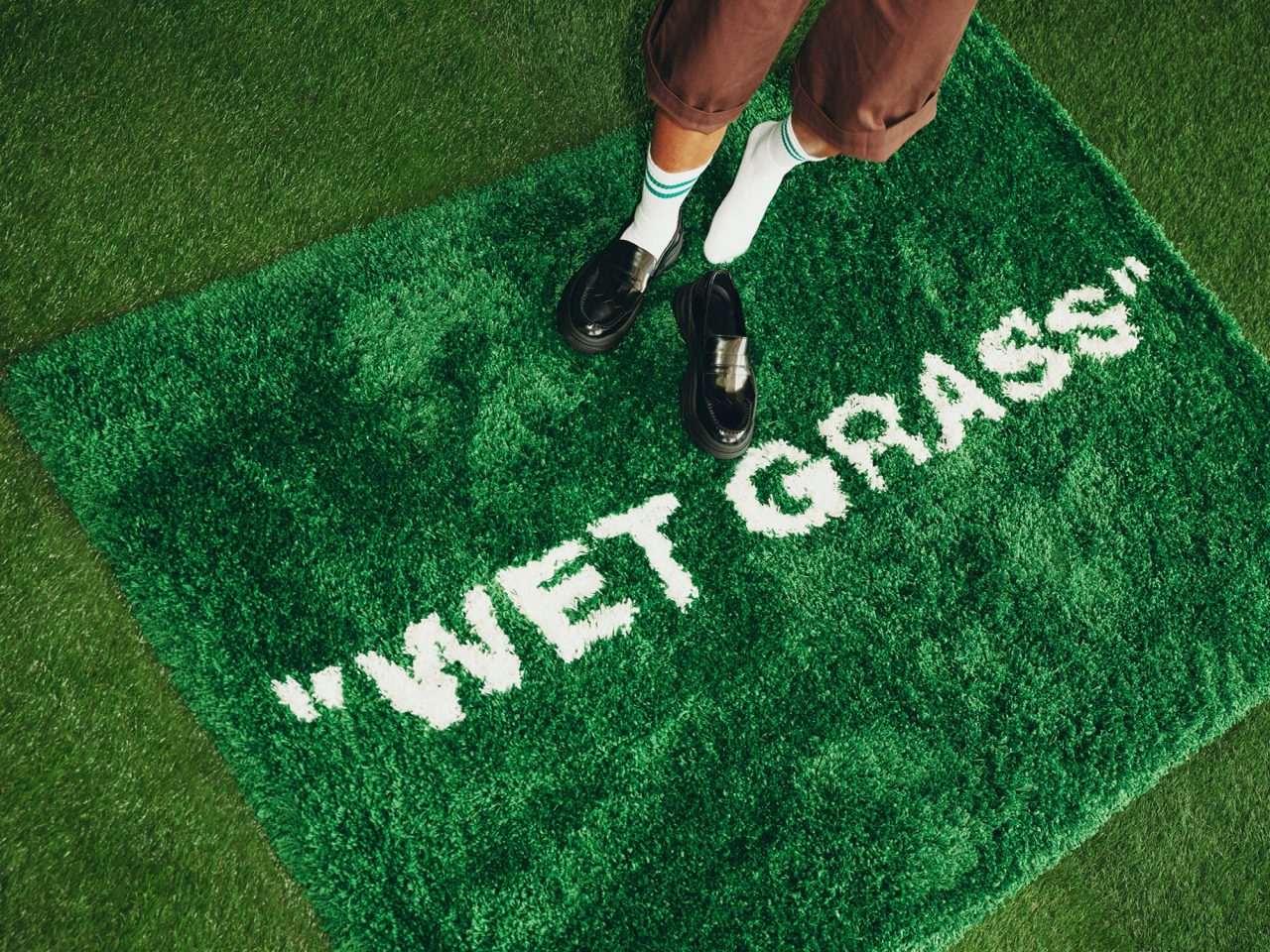 Person in shorts, white knee socks and black shoes standing on a green rya rug with the white text: WET GRASS.