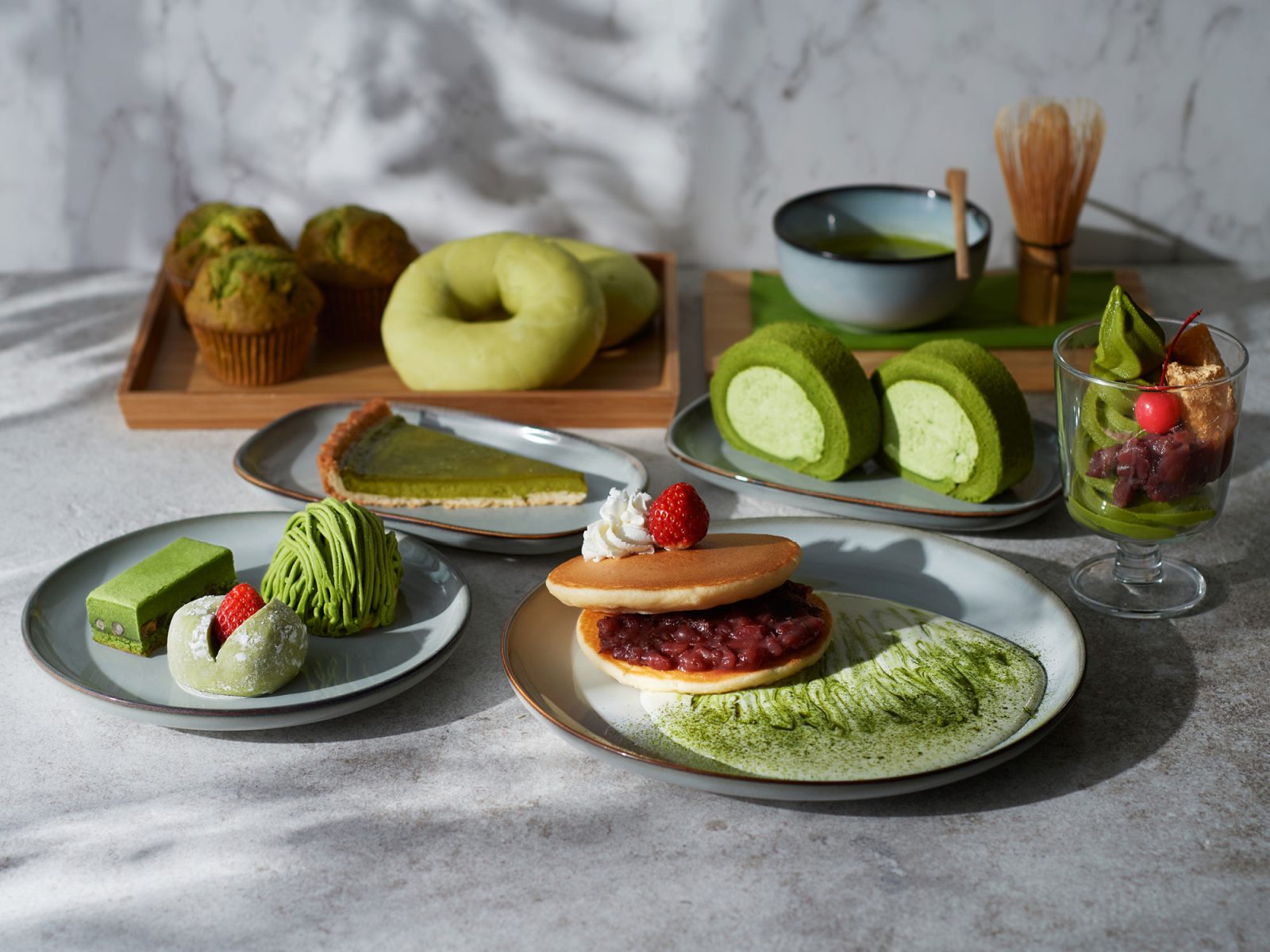 Set table with sweets, everything from doughnuts to slices of cake, a traditional matcha whisk and a bowl of green tea.