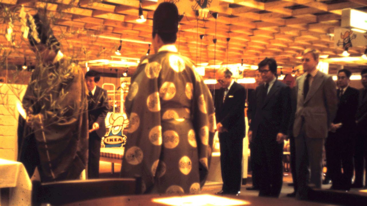 Two Shinto priests performing ceremony in shop area, group of men in suits standing respectfully to the side.