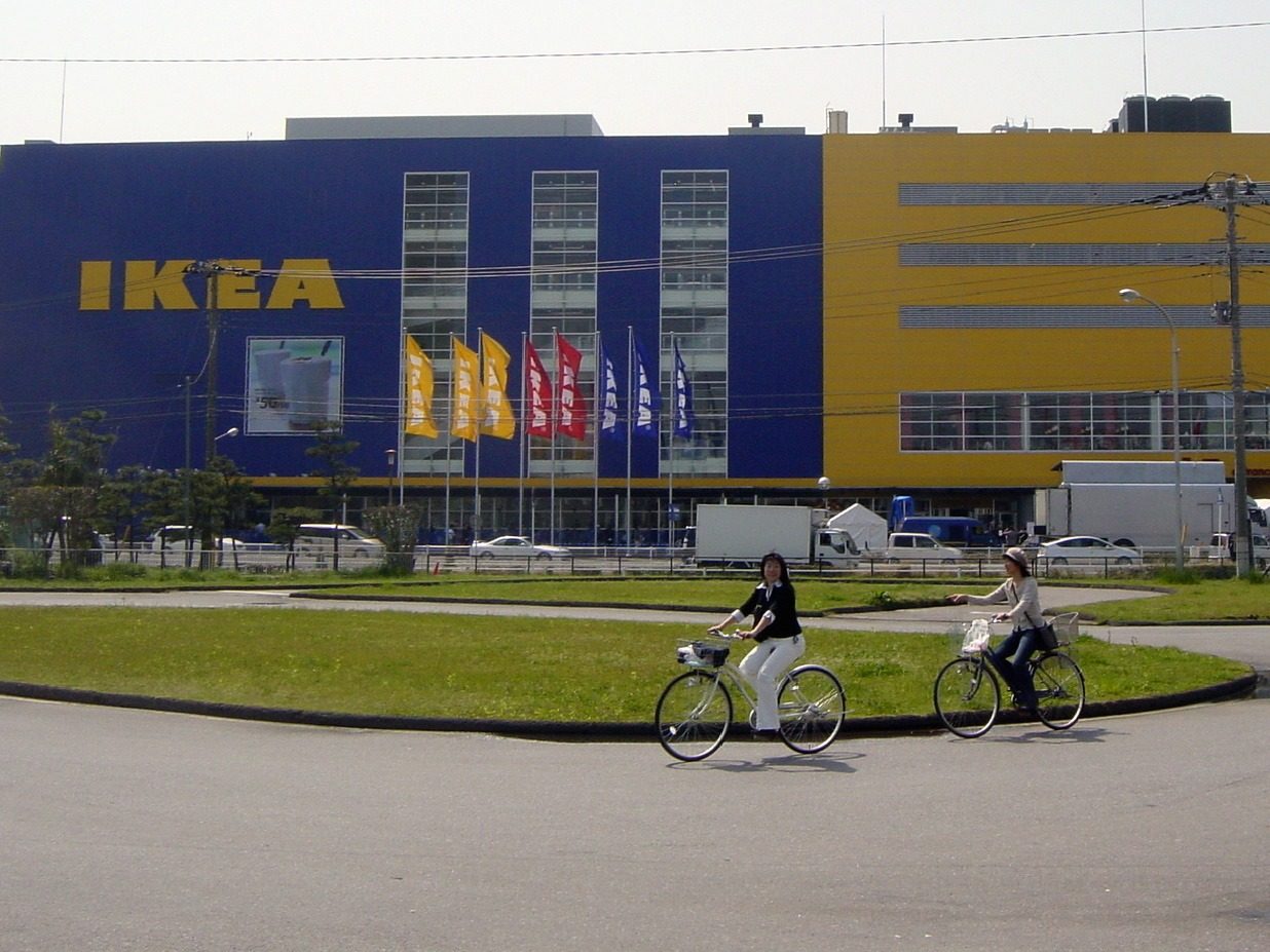 Two women cycling in front of a large blue and yellow IKEA warehouse.