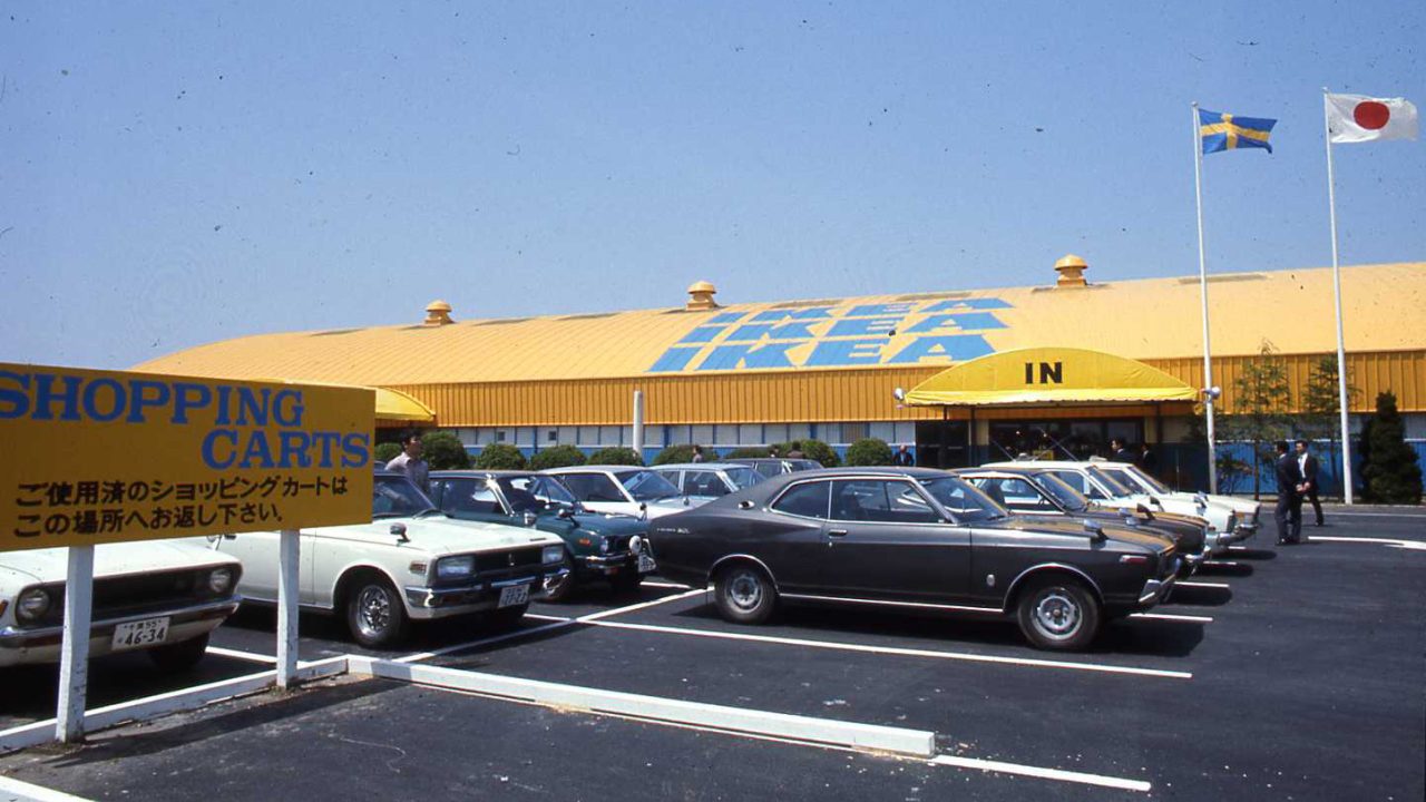 1970-talsbilar parkerade framför en blå och gul IKEA butik där man hissat en svensk och en japansk flagga.