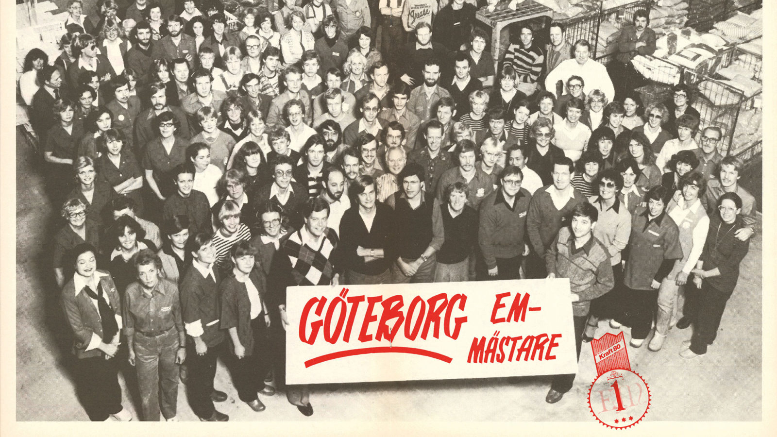 Svartvitt gruppfoto av en stor samling personal på IKEA i 1980-talskläder bakom skylt med text 