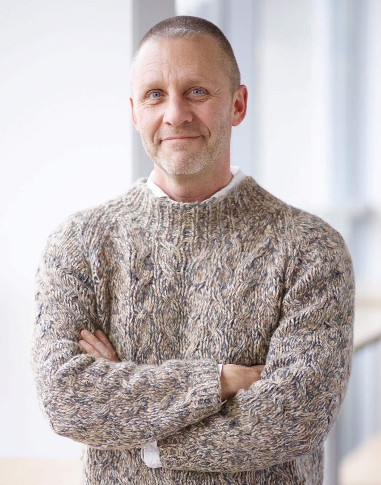 Man som ler, med korslagda armar. Han har kort grått hår, stickad gråbrun tröja över vit skjorta och svarta byxor.