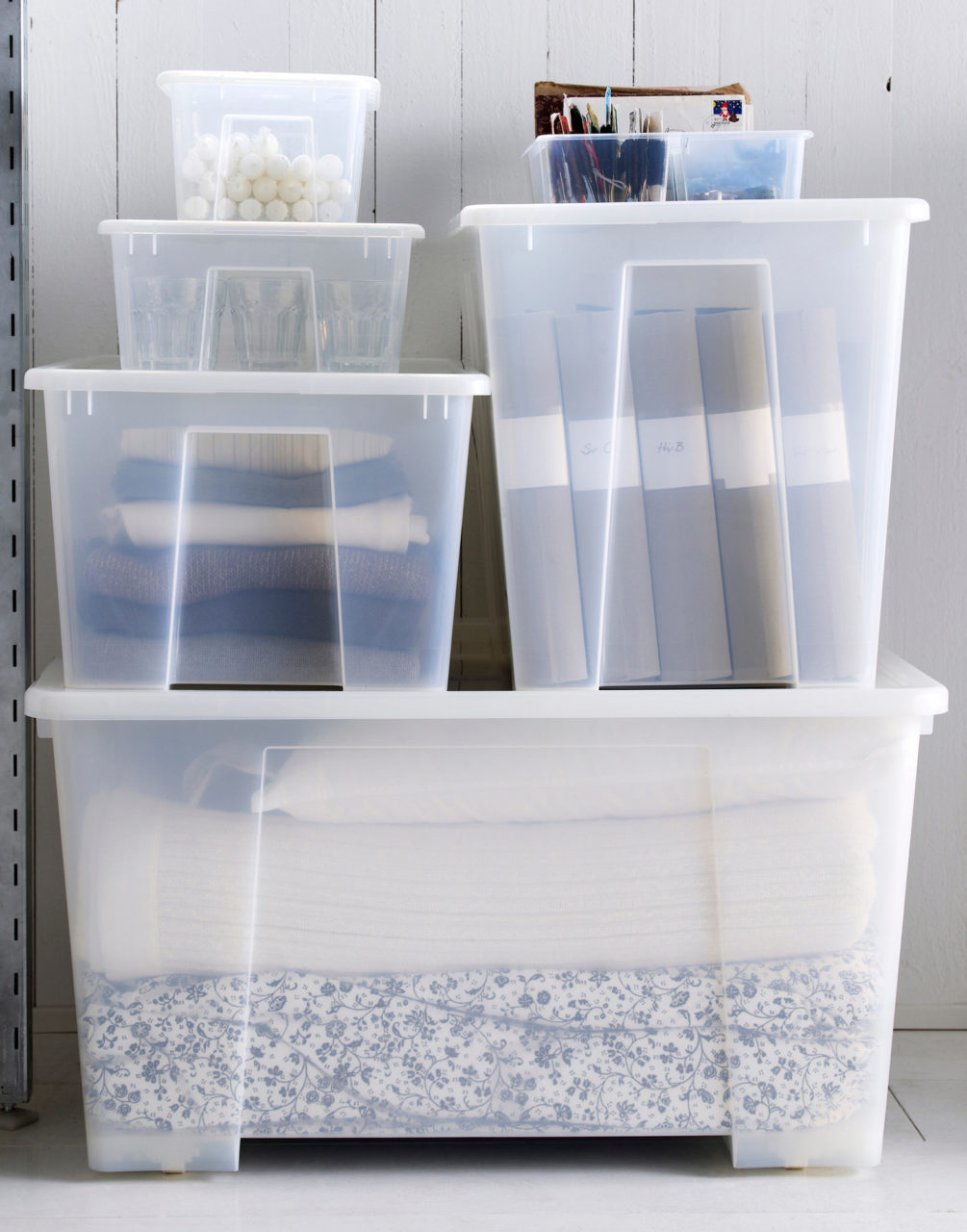 Contenitori in plastica di misure diverse impilati. Al loro interno, tessili per il letto, vestiti, raccoglitori e candele.