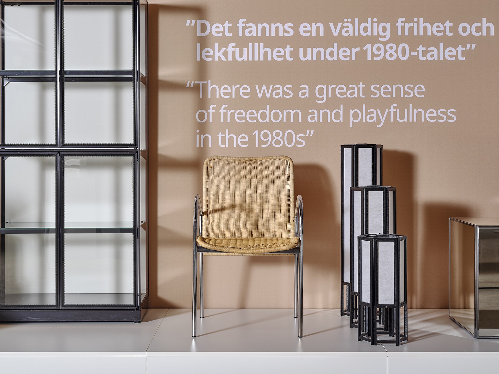 Black glass cabinet, rattan chair and other furnishings displayed on a podium against a beige wall with a close-up of a wedge dowel.