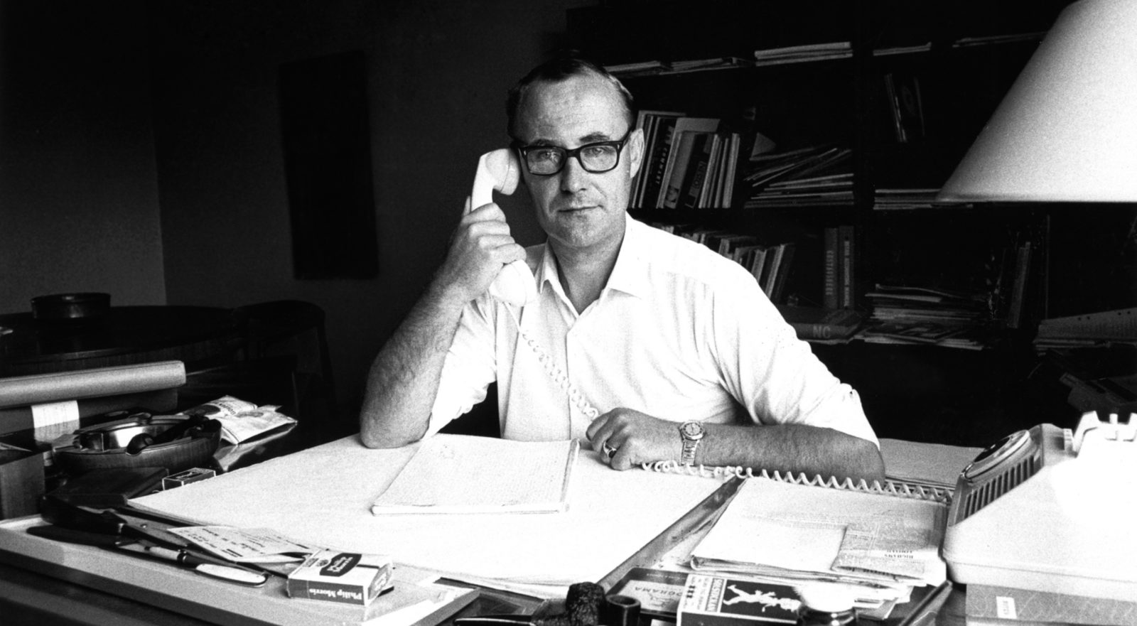 A black and white photo of Ingvar Kamprad at his desk, taking notes while talking on the phone.