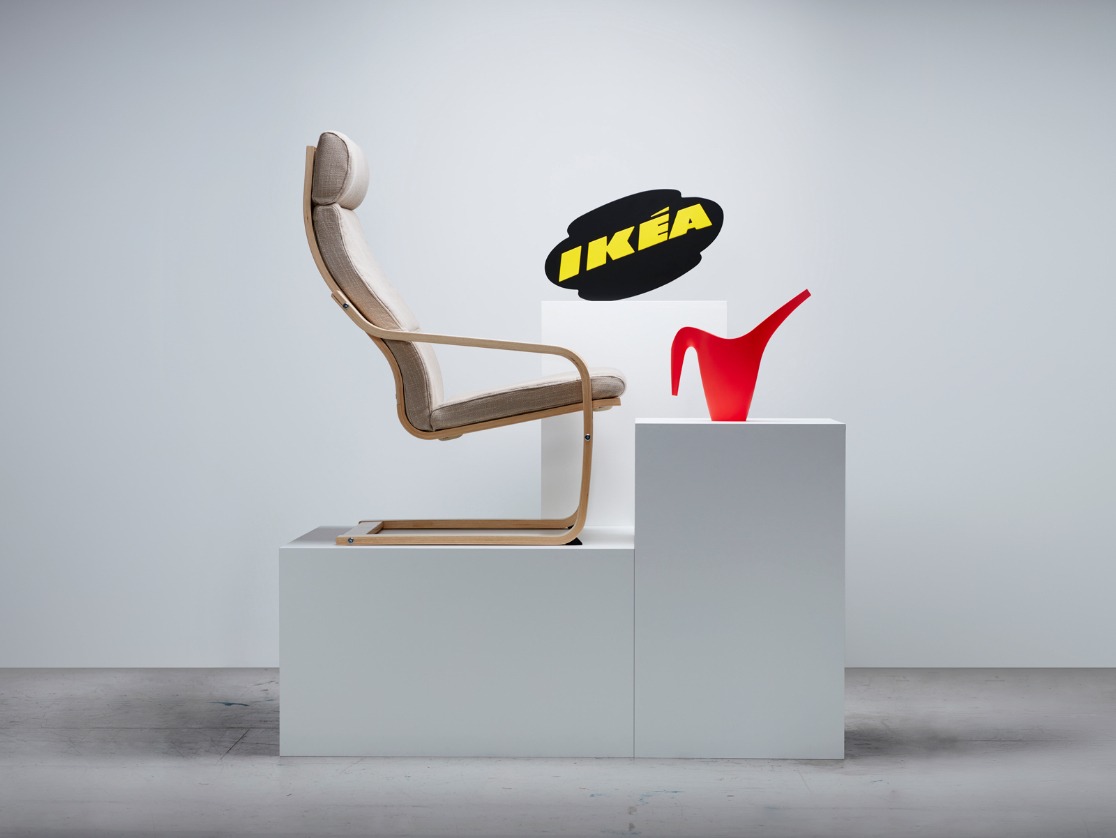A podium with a beige POÄNG chair, a red IKEA PS 2002 watering can, and a black and yellow IKEA logo from the past.