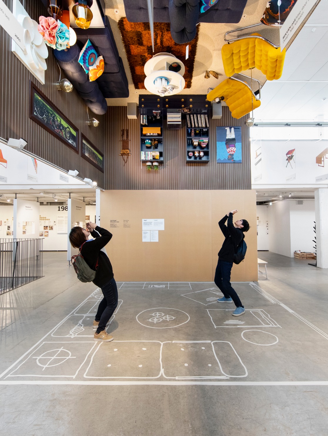 IKEA Museum visitors taking photos of an upside-down Seventies room with yellow, blue and brown furniture attached to the ceiling.