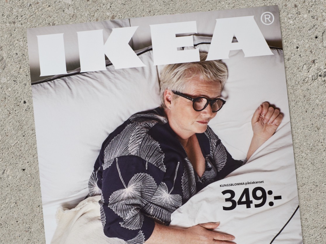 A modern-style IKEA catalogue cover with an IKEA Museum visitor lying in a bed made with a white quilt cover and cushions.