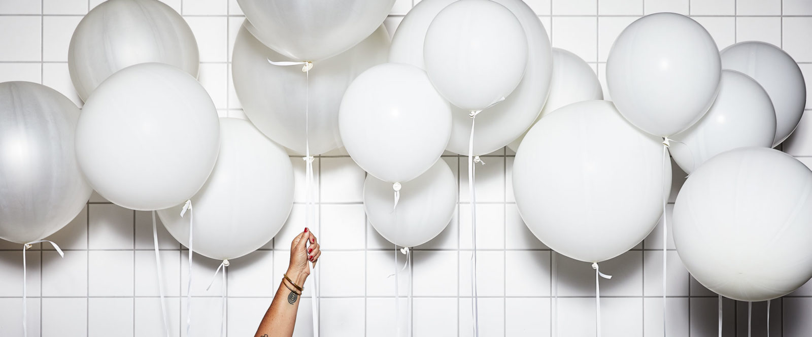 Several white, floating balloons with white strings and a hand holding one of them in front of a white, tiled wall.