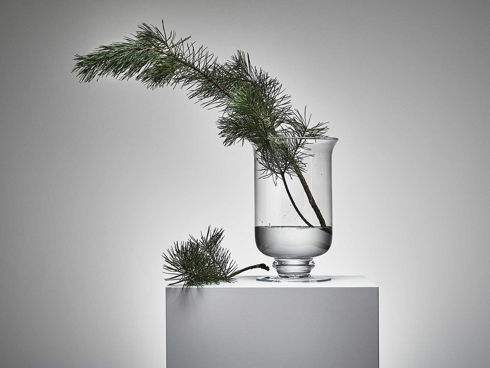 Vase or lantern in mouth-blown glass adorned with a spruce twig, standing on a podium.