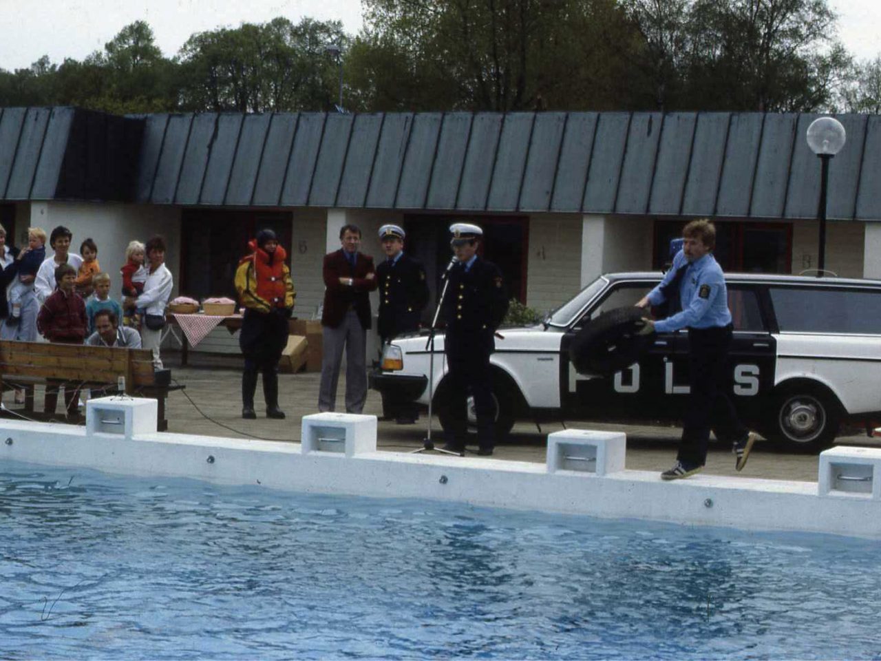 An audience and several police and a police car at the edge of a pool, a policeman throws a life jacket into pool.