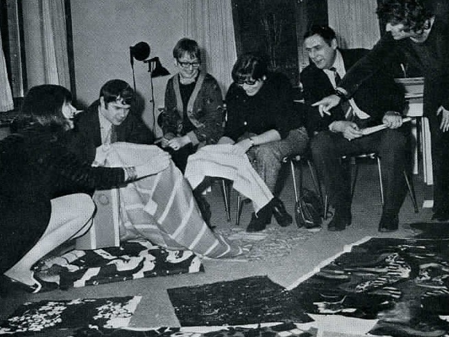 Group of smiling people in 1960s style clothing looking at striped fabrics.