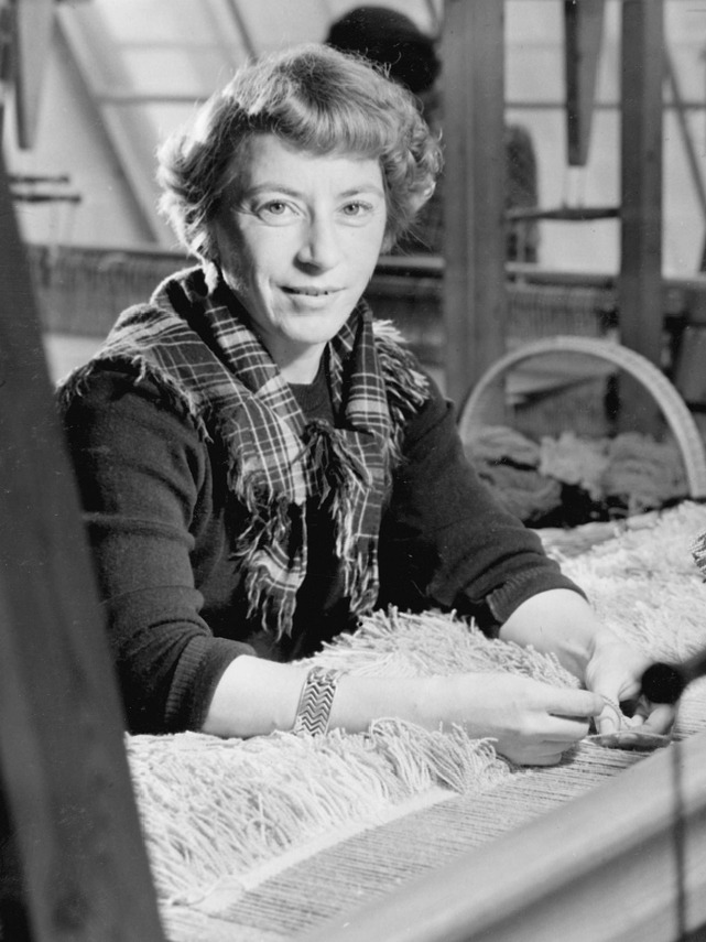 Viola Gråsten, a blonde woman in her 30s, sits at a loom dressed in a sweater, checkered shawl around her neck.