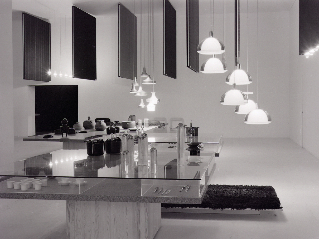 Exhibition with stylish design, large table in glass and wood, chrome lamps hang from the ceiling.