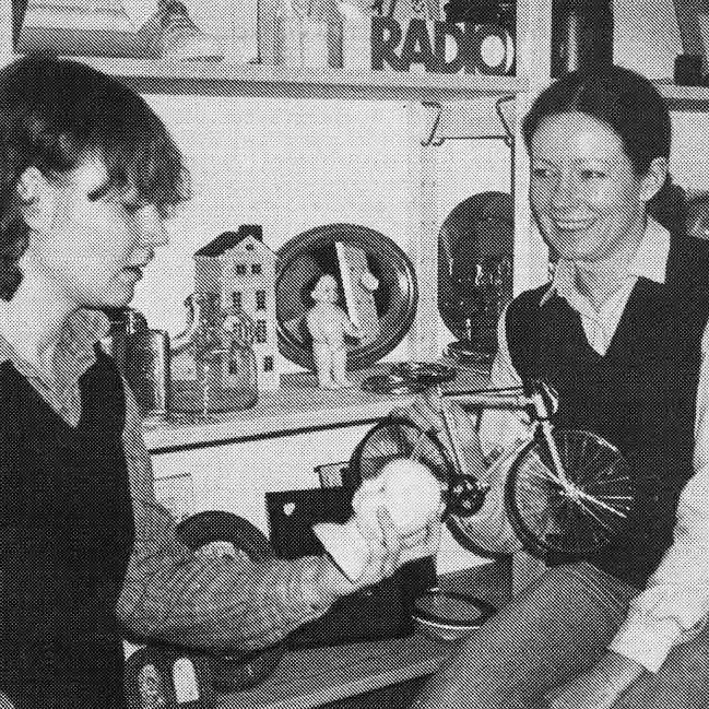 Two women in front of a bookshelf with decorative items; one holds a small white sculpture.