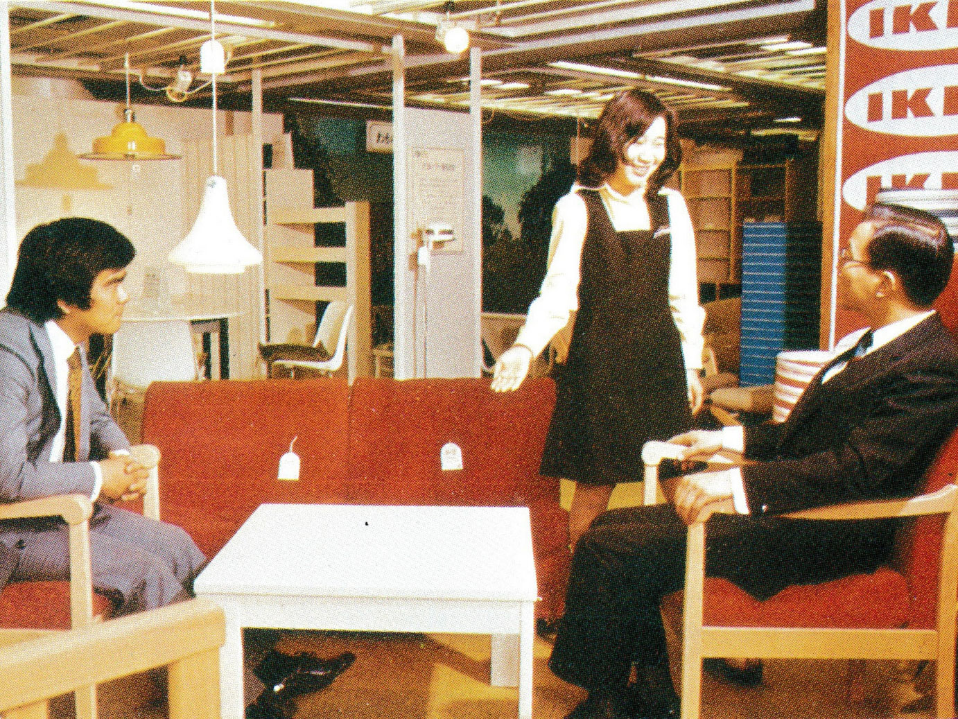 アームチェアに座るスーツ姿の男性2人と女性コワーカー1人。1970年代の日本の百貨店内に展示されているイケアの家具。