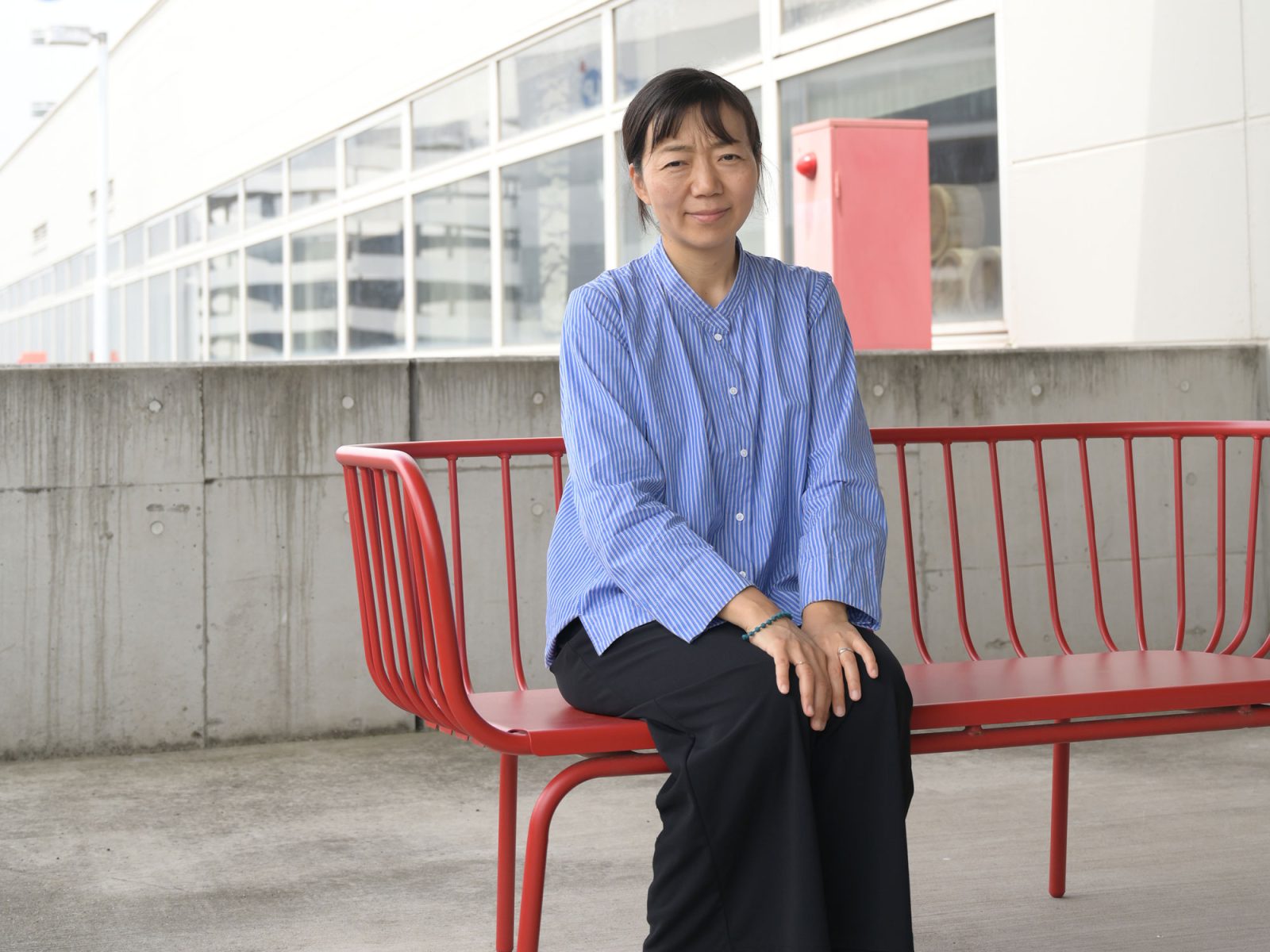 Dark-haired woman in blue blouse sitting on a red metal sofa, Aya Kosai.
