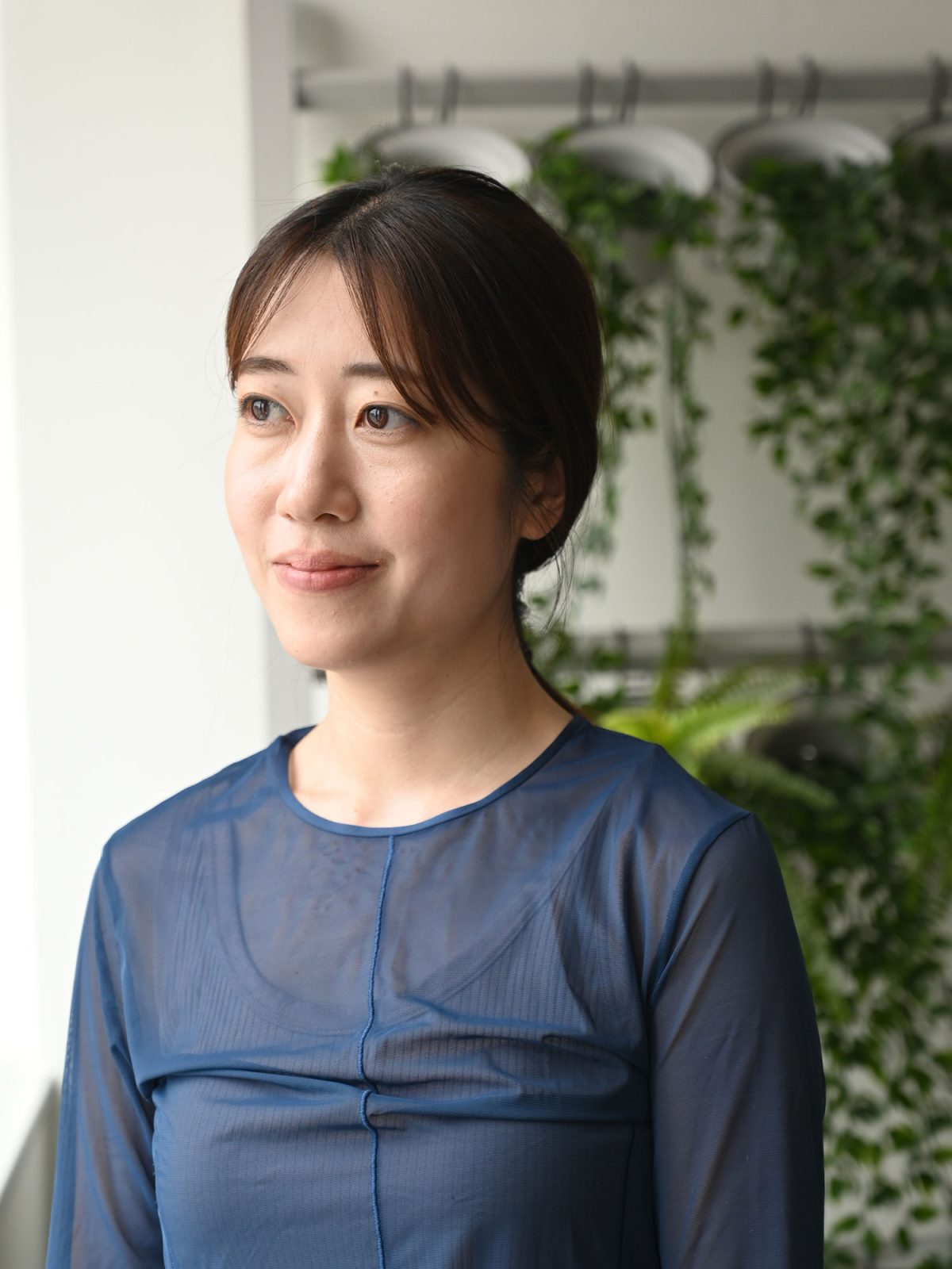 Dark-haired woman in blue blouse, Mai Tanigawa, standing in front of a wall with green plants.