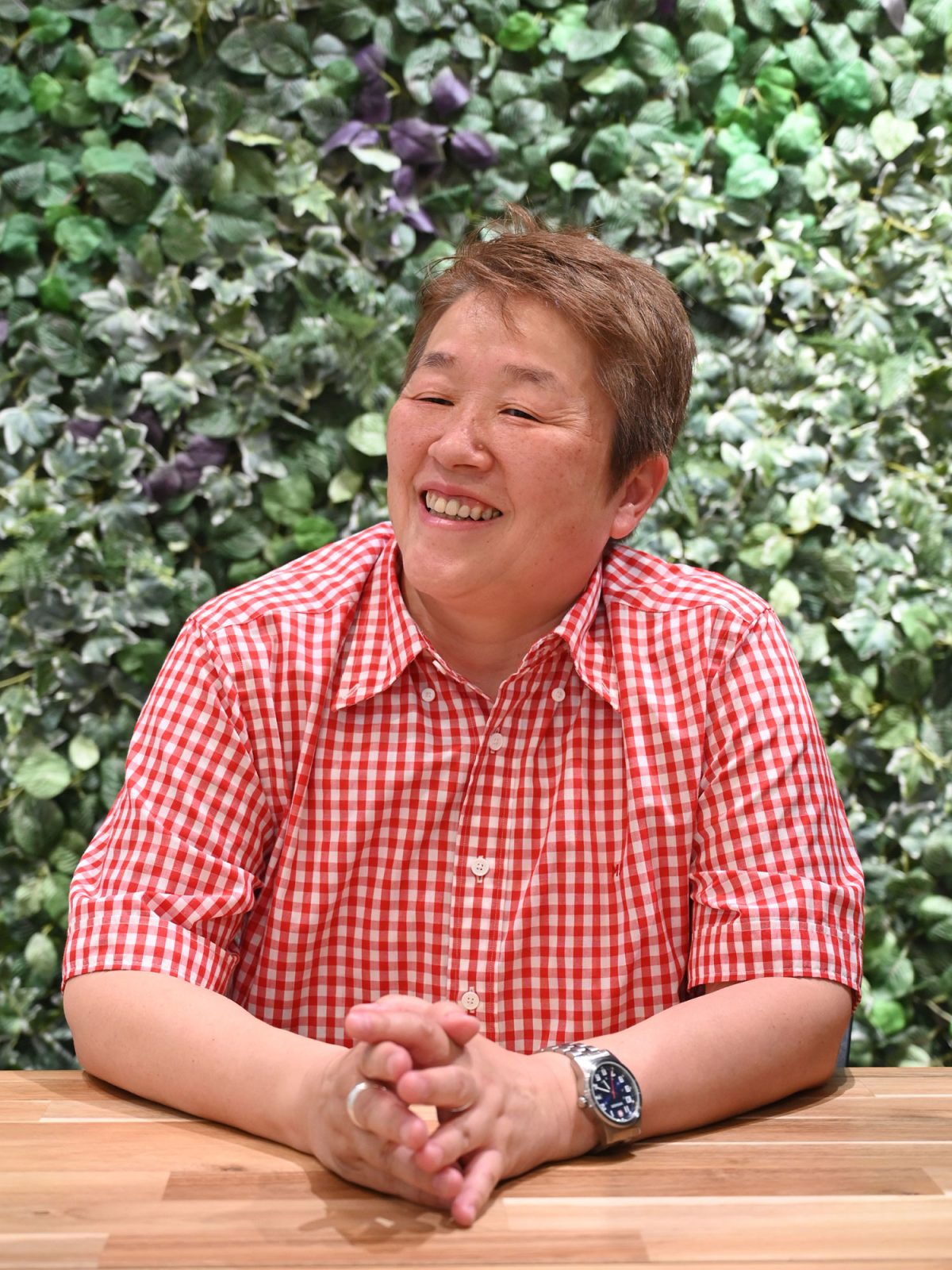 Smiling dark blonde woman, Tamae Asayama, in red and white check shirt sitting in front of a wall with green plants.