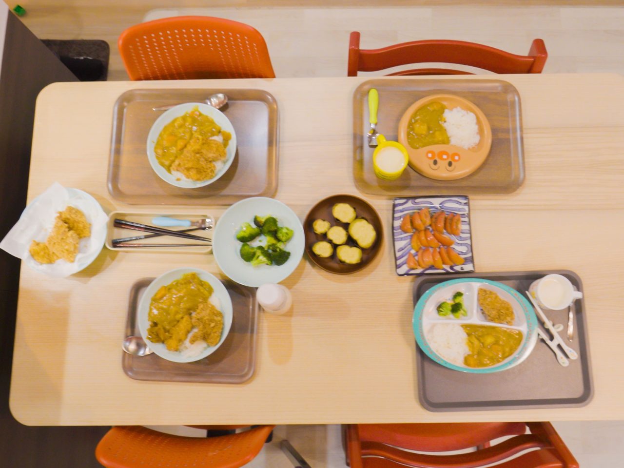 Dining table viewed from above, four trays with food served and some smaller bowls with vegetables.