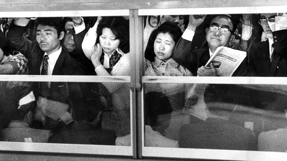 非常に混雑している日本の地下鉄車内の白黒写真。疲れた表情の男性や女性が写っている。