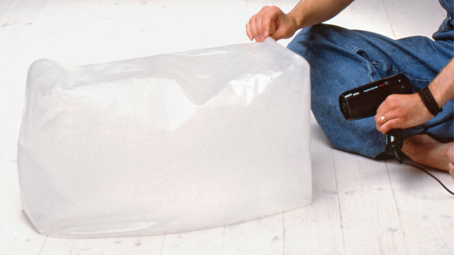 Young man in dungarees sitting on the floor in a photo studio, inflating a plastic pillow with a hair dryer.