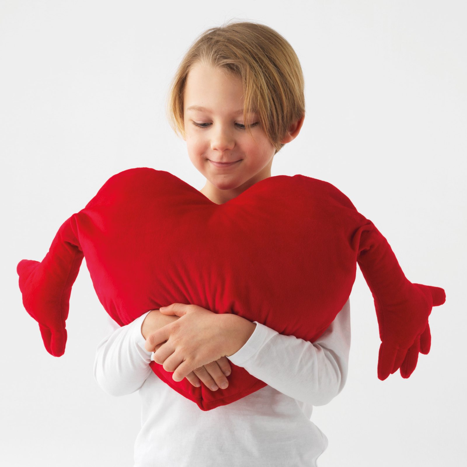 Blonde child in white T-shirt hugs a soft, heart-shaped red pillow, FAMNIG, which has outstretched arms.