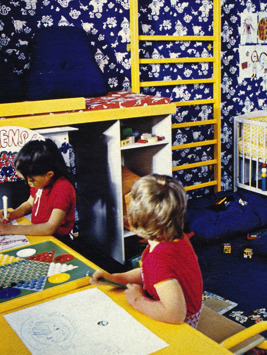 Två barn i 1970-talskläder sitter vid ett skrivbord och ritar och målar, i bakgrunden syns en ribbstol på väggen.