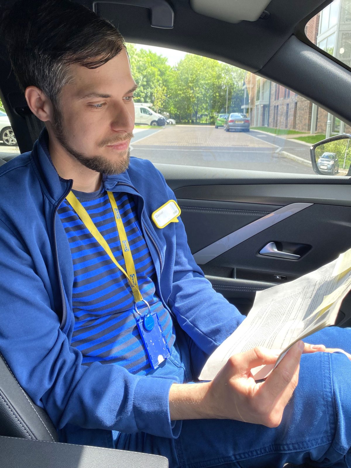 A young dark-haired man, Daniel Farago, dressed in IKEA work clothes, sits in a car studying some papers.