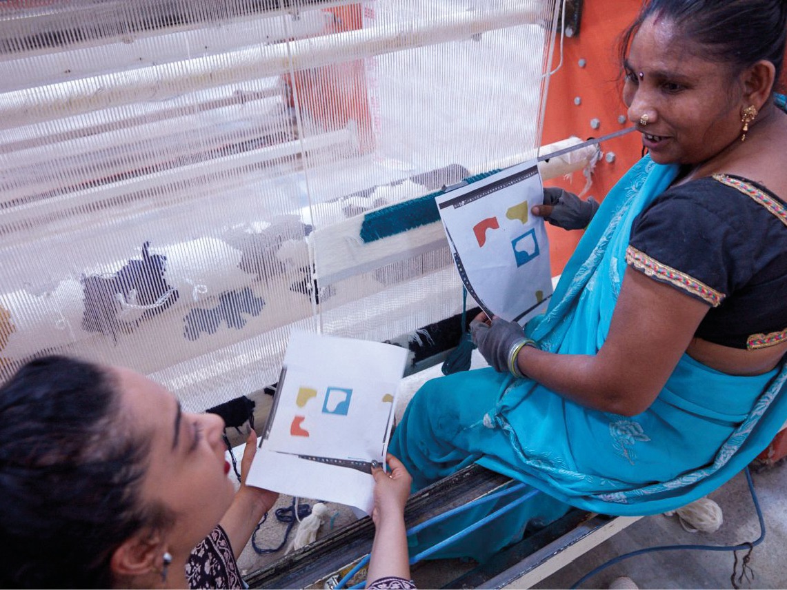 Dark-haired woman, Akanksha Deo, shows a sketch to a woman in a blue sari, who is sitting at a loom.
