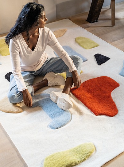 Dark-haired woman in white sweater sits on large rug with 3D patterns in different scales and colours.