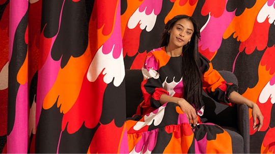 Darkhaired young woman in dress with bright-coloured pattern in orange, white, pink, red and black.