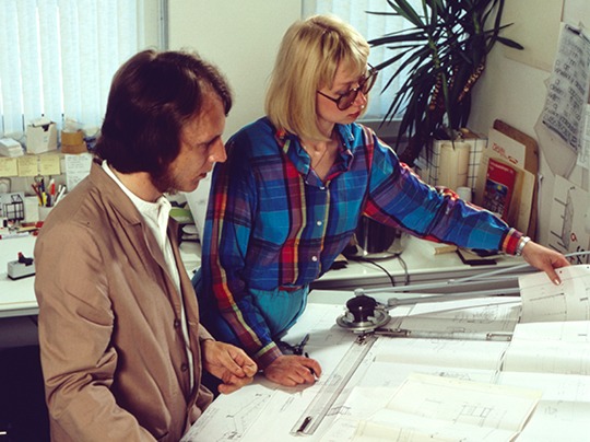 Young man and woman working at drawing table, wearing 1970s clothing.
