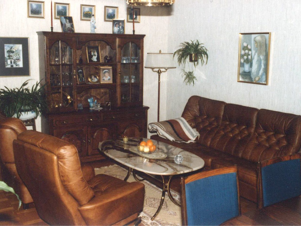 A 1980s-style living room features leather furniture, a display cabinet, and a small crystal chandelier.