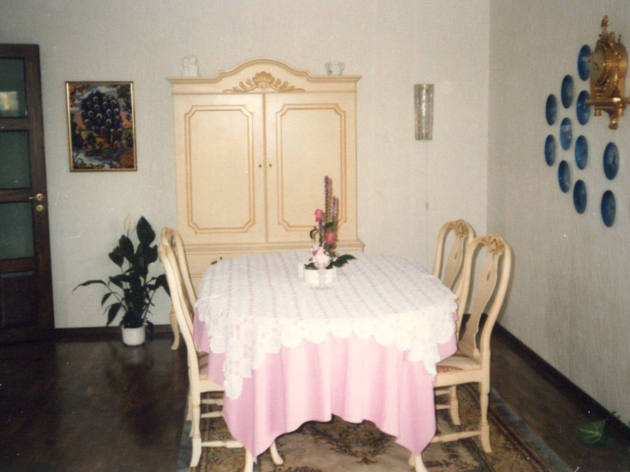 A dining set made of white wood has a white tablecloth, with old-style oval-framed pictures hanging on the wall.