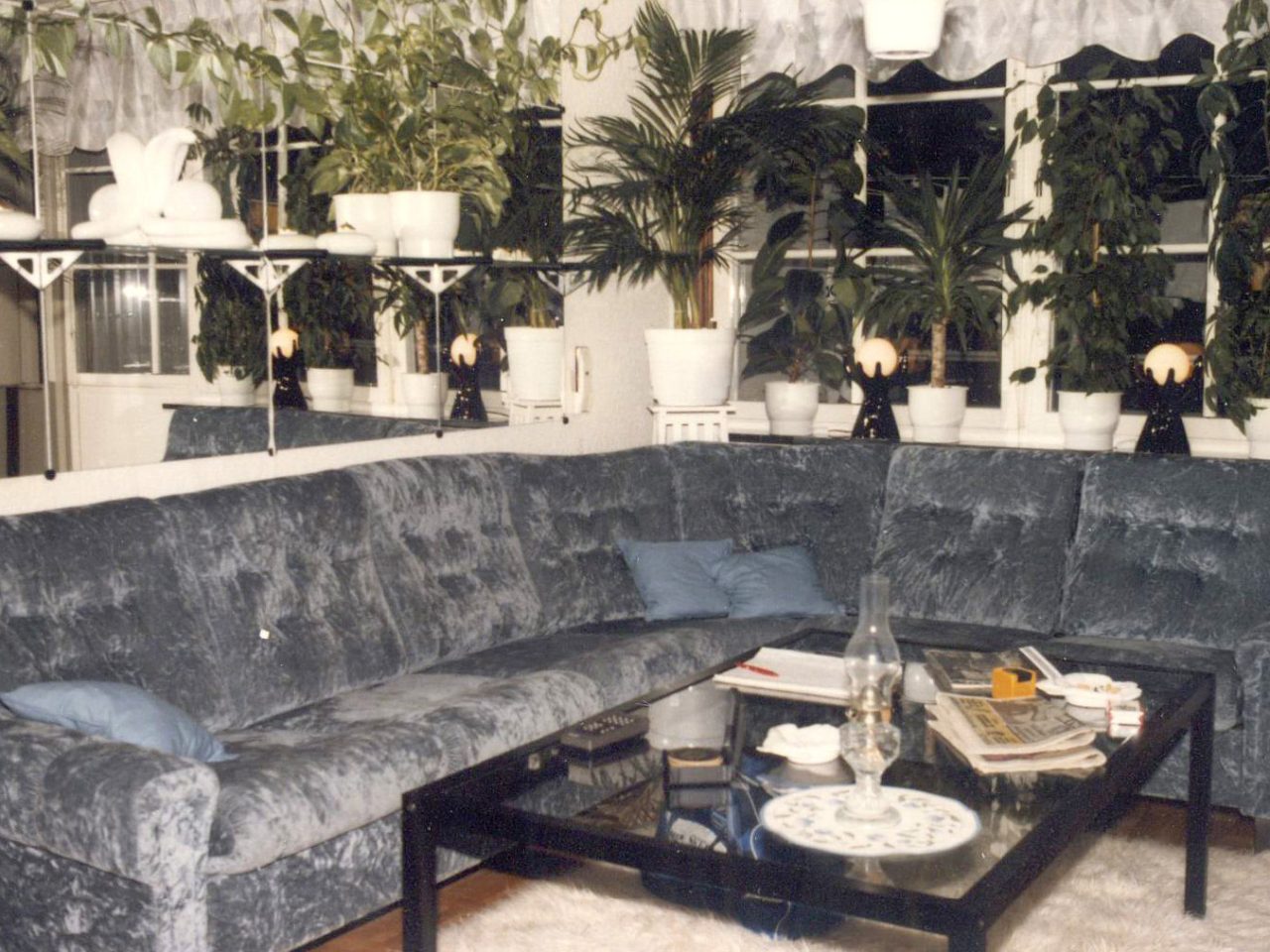A living room with a large grey velvet corner sofa under a mirrored wall, with many green plants.
