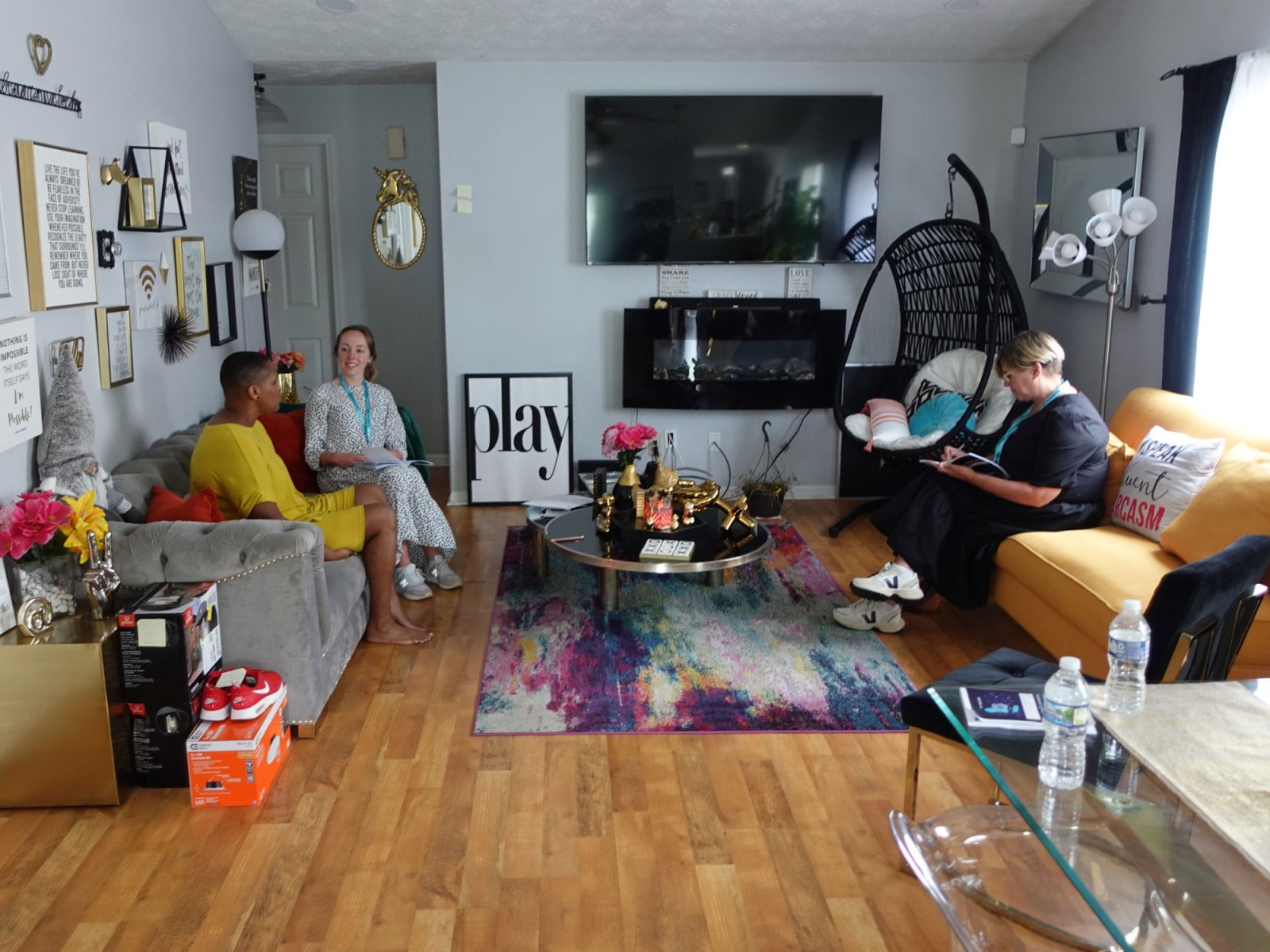 Three women sit in two sofas in a well-filled living room, one of them taking notes in a notebook.