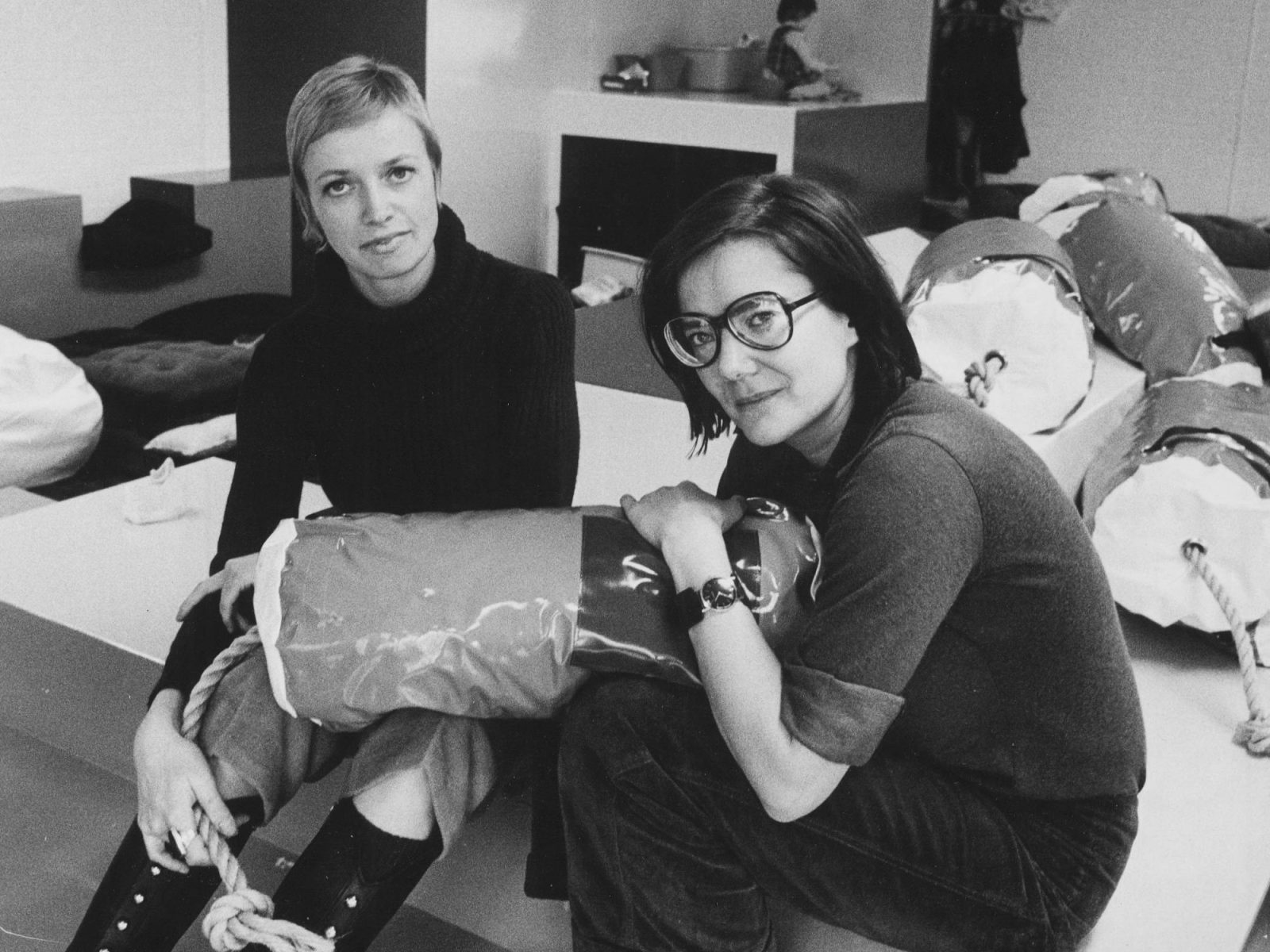 Two young women sit in a large playroom, one holding a soft oblong pillow.