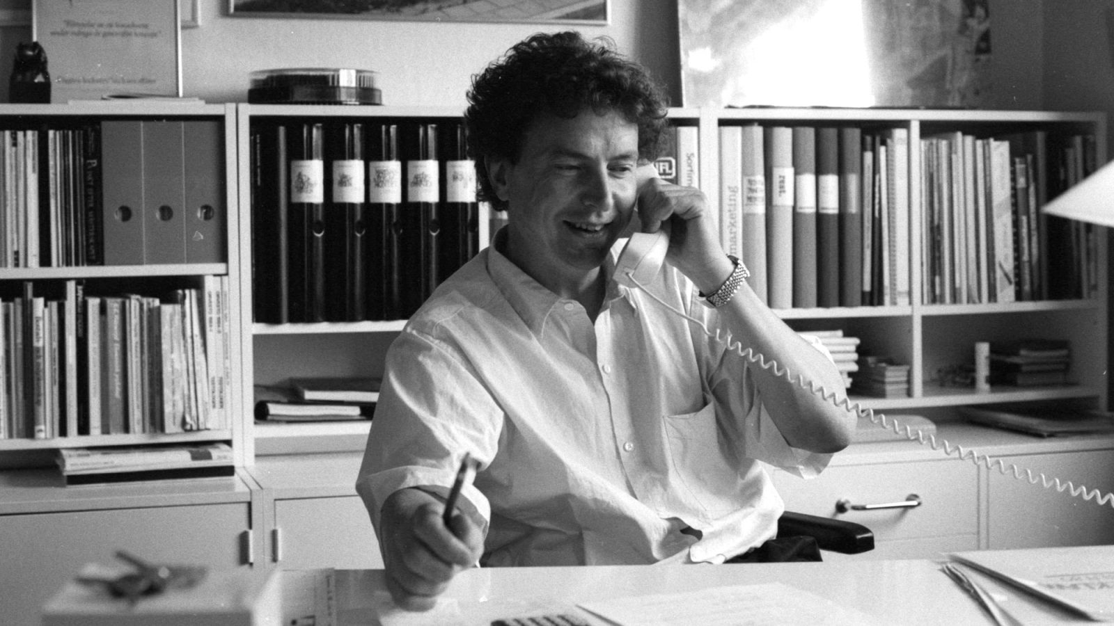 A man in a white shirt, Lennart Ekmark sits at a desk talking on an old phone, with binders visible in the background.
