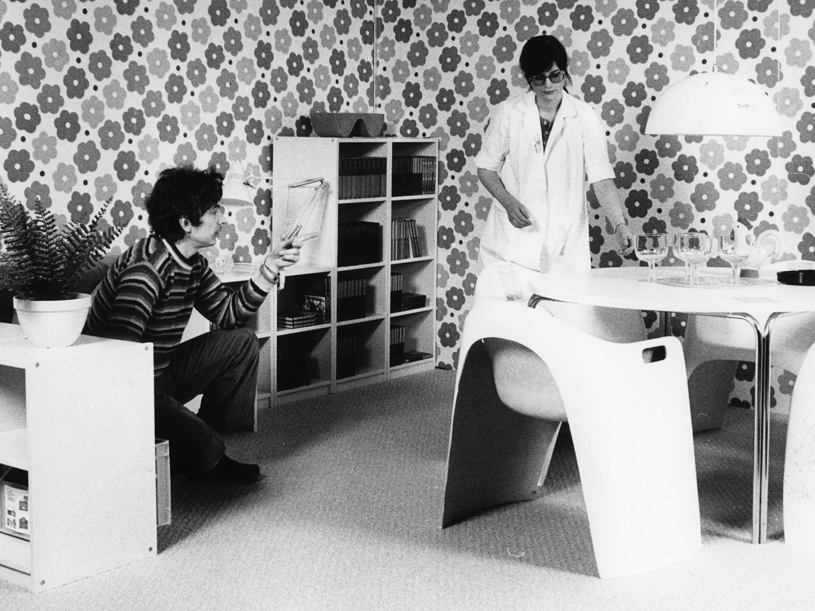 Two young people in 1960s clothes sit in a room furnished with black-and-white furniture trendy at the time.