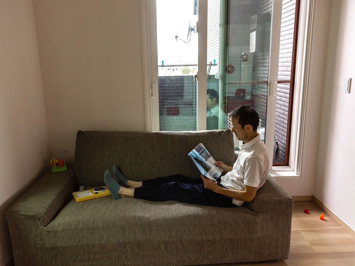 A man with a Japanese appearance lounges on a small brown sofa reading a newspaper.