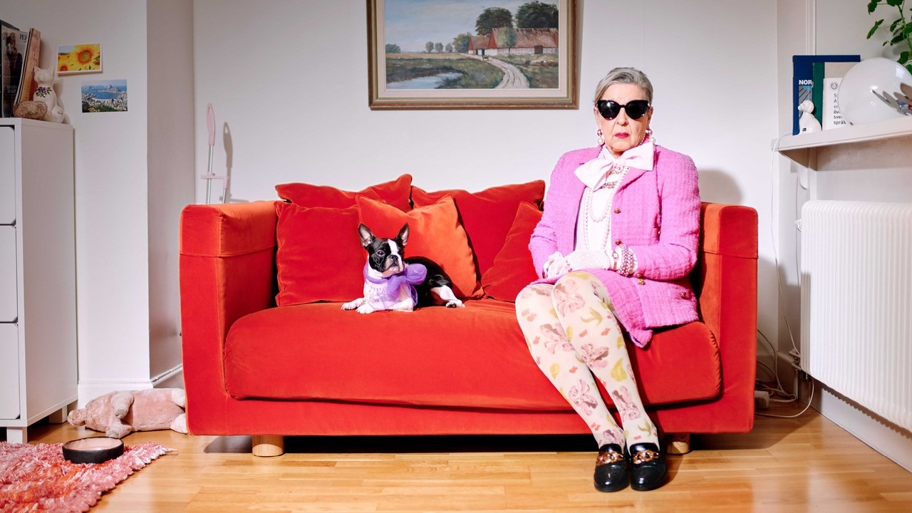 An older woman wearing sunglasses, a pink jacket, and floral tights sits on an orange sofa. Beside her lies a dog with a purple bow around its neck. Above them hangs a painting.