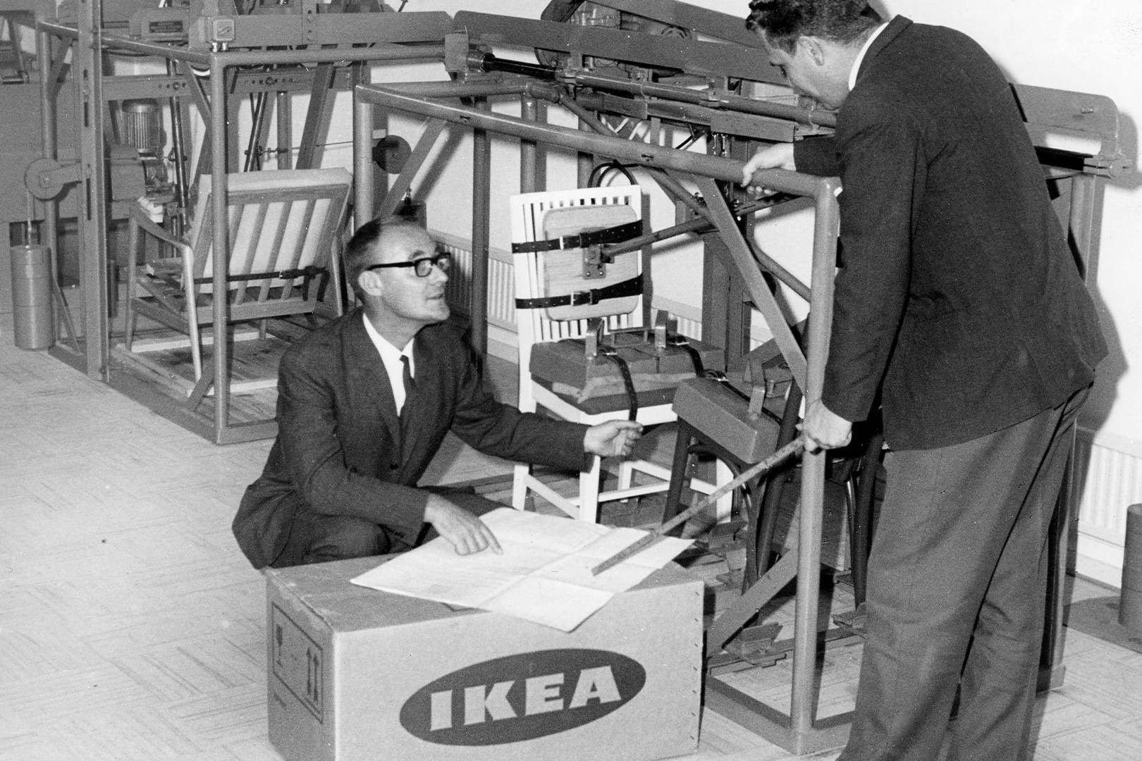 Ingvar Kamprad testing a chair in a factory with a co-worker, with an IKEA box and machinery in the background